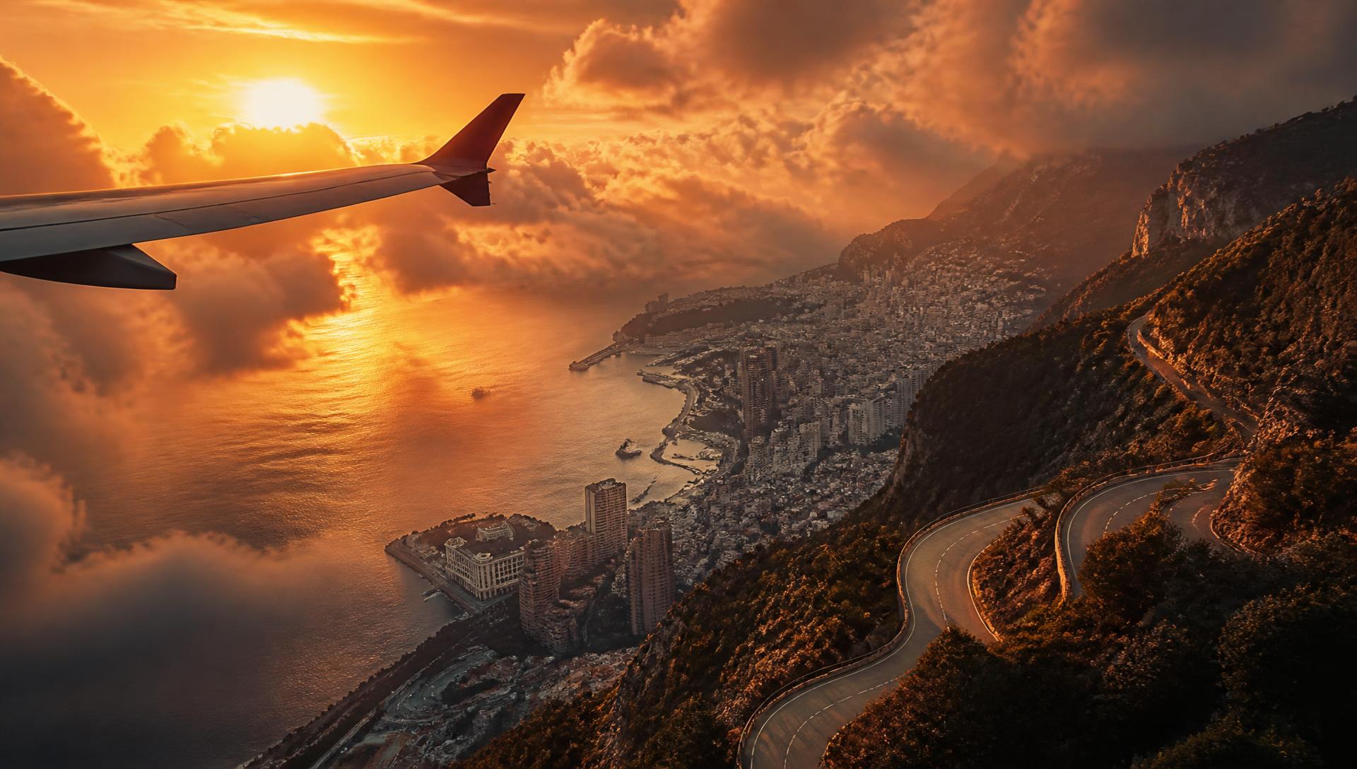 Aircraft wing above golden sunset clouds over a coastal city and mountain road
